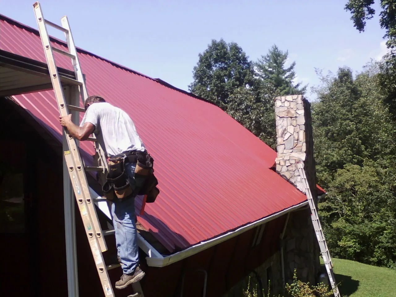 Roofer on a ladder beside a red metal roof and stone chimney — Soffit & Fascia Repair by Roofing Brothers in Peachtree City